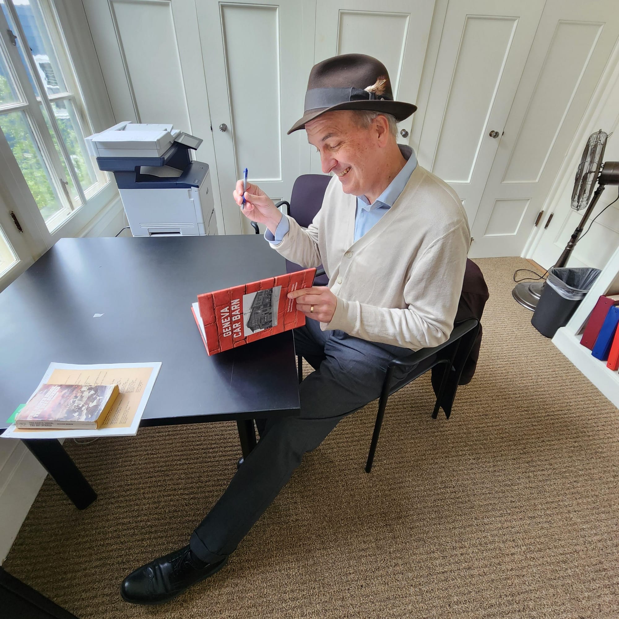 Man wearing beige cardigan and gray fedora signs a copy of a Geneva Car Barn book.