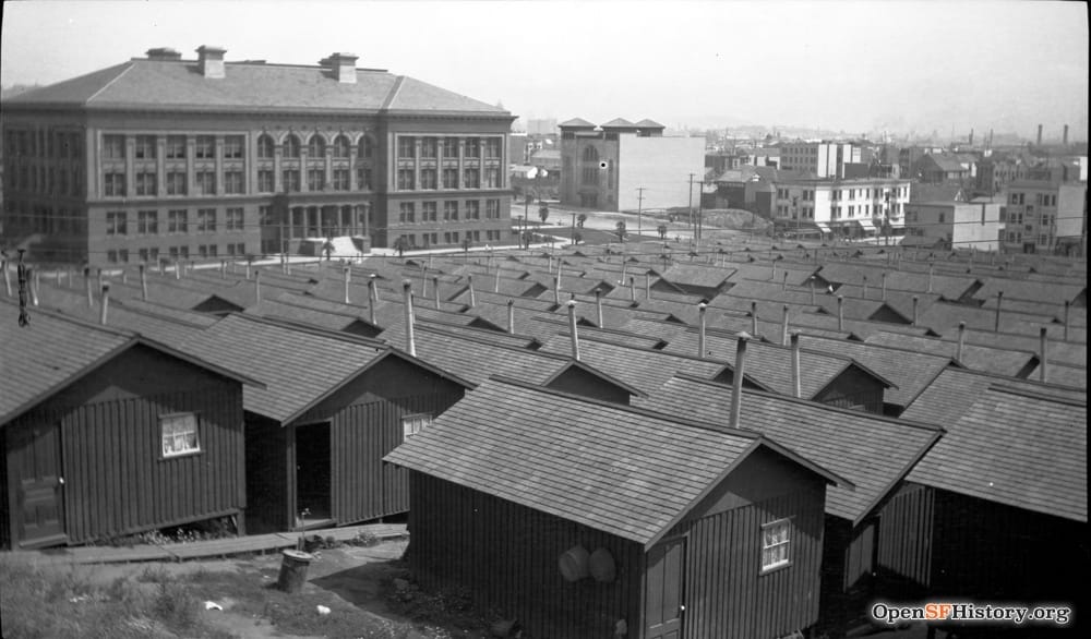 Black and white photograph looking upon a sea of one-room earthquake cottages in a park, with large four-story school building in the background.