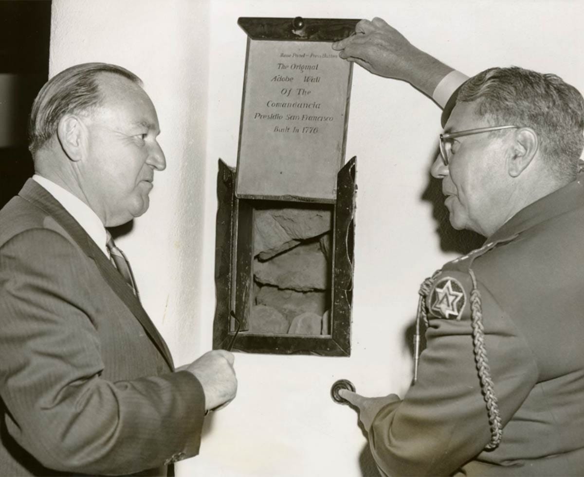 Two men, one wearing a suit and one a lt. uniform, look through a square opening of plaster to pieces of adobe bricks.