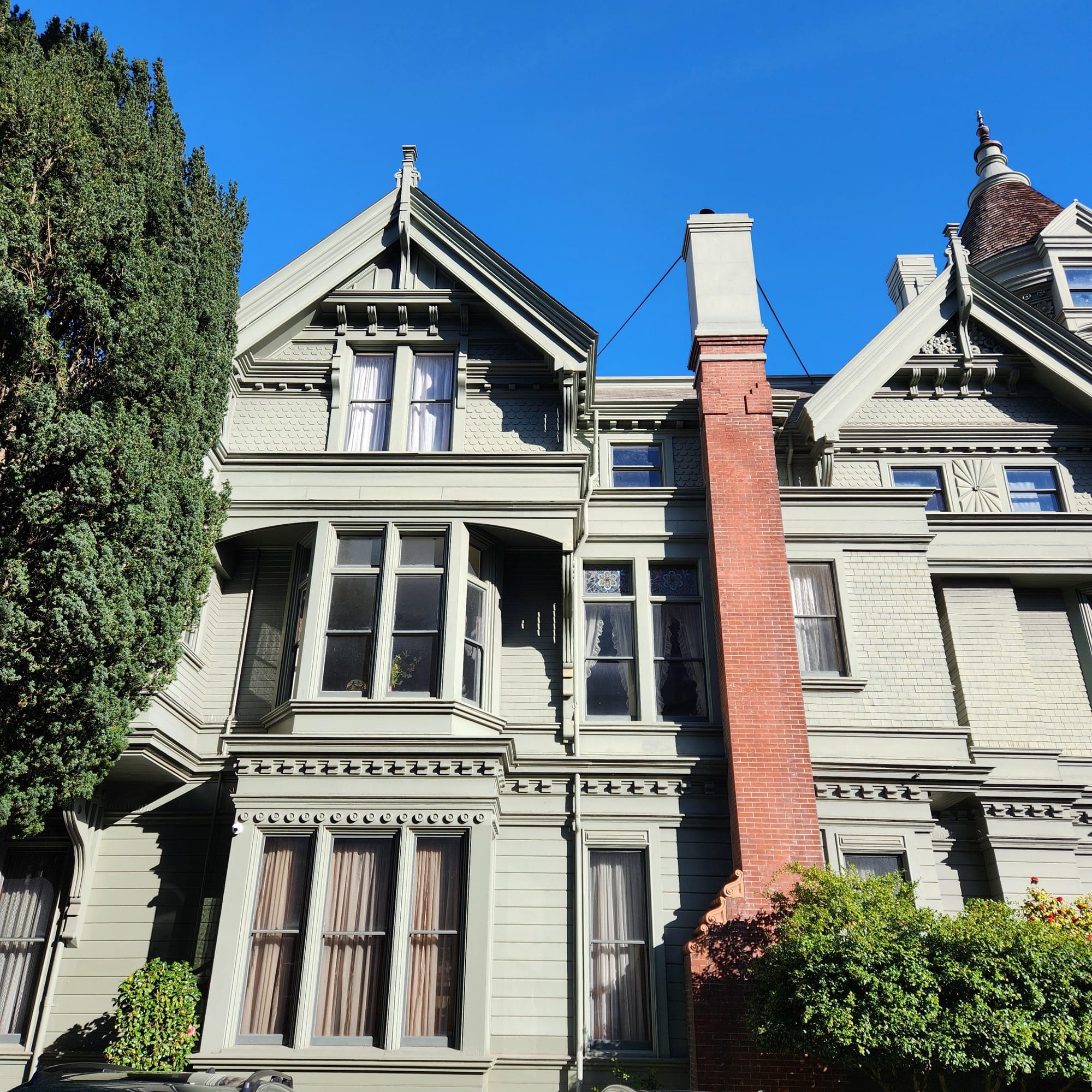 Side view of three floors of a green-gray Queen Anne Victorian home, with a terra cotta chimney 