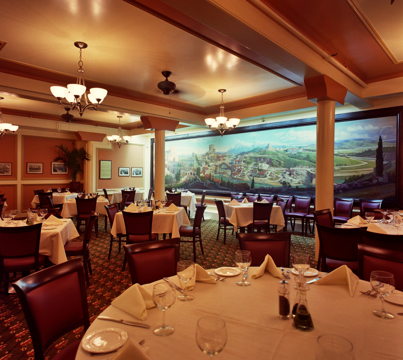 Photograph of the inside of a restaurant, featuring tables with white tablecloths and a long horizonal wall mural of a northern italian scene.
