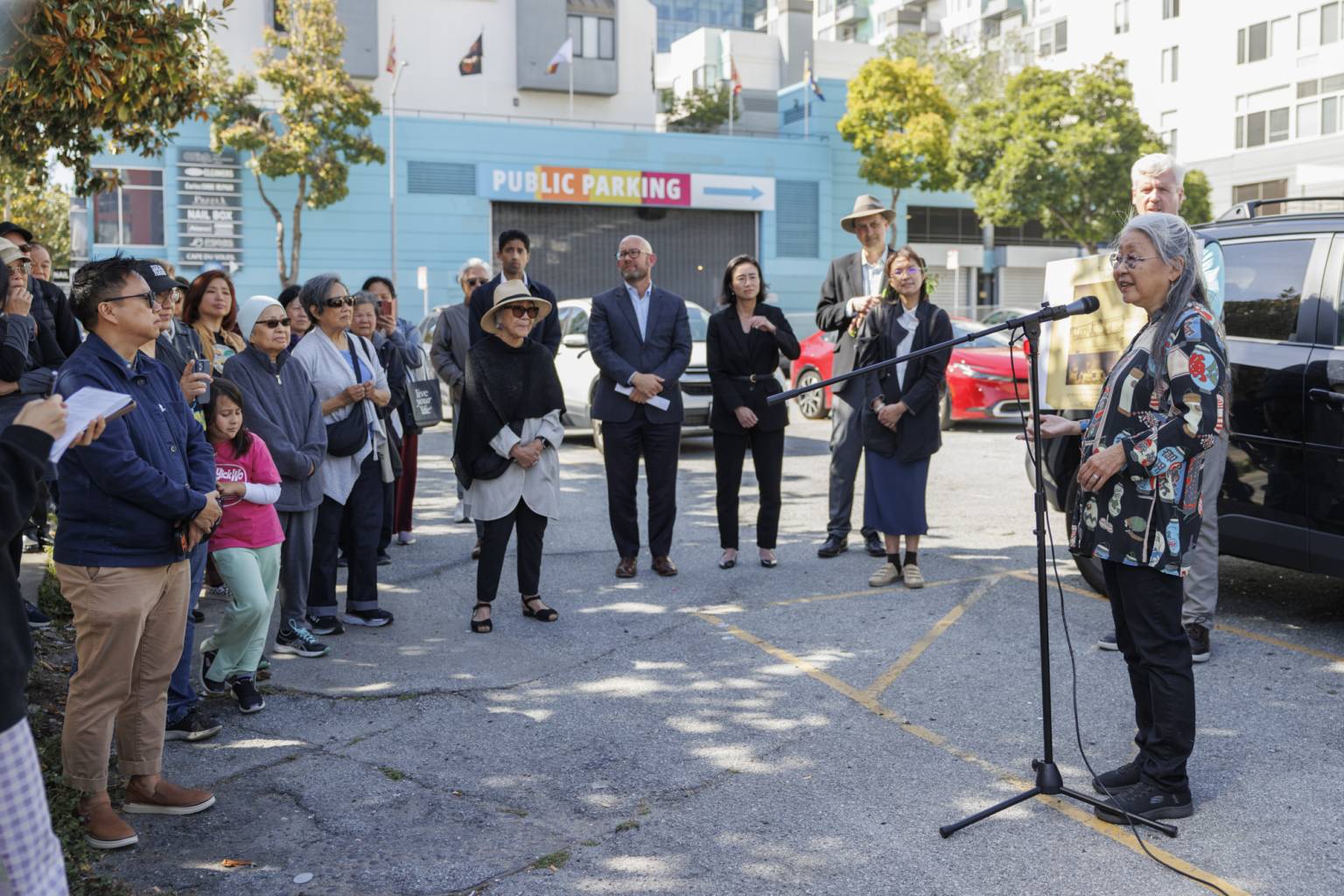 A woman with long gray hair and glasses speaks into a standing mic to a small crowd in a parking lot.