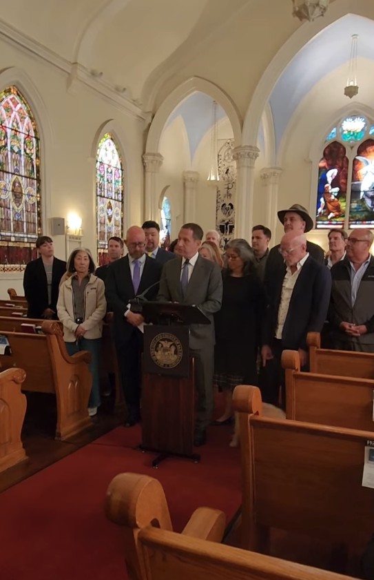 Screenshot from a video featuring a man in gray suit and blue tie standing behind a podiu speaking, with group of people behind him.