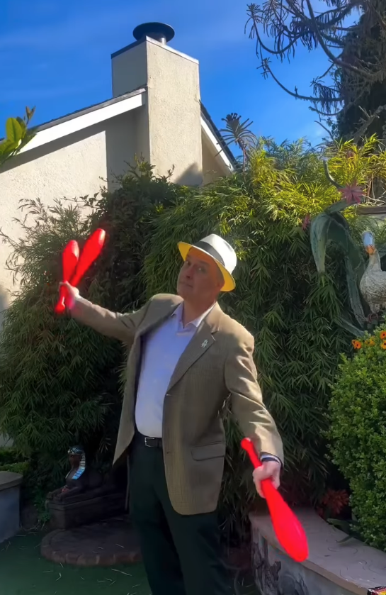 Vide screenshot of a man wearing a white fedora and holding three red juggling pins .