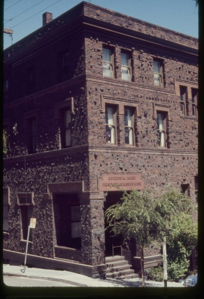 Exterior view of corner of three story brown clinker brick building with street tree on sidewalk in front.
