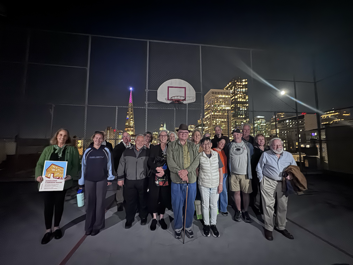 A group of 17 people pose for a photo on an outdoor basketball court with SF city skyline in the distance.