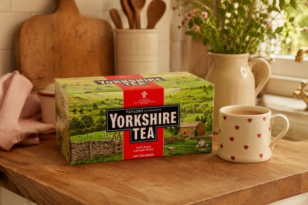 A box of Yorkshire Tea on a wooden kitchen worktop beside a mug of tea, with a jug of flowers, chopping boards and a pot of wooden spoons by the window.