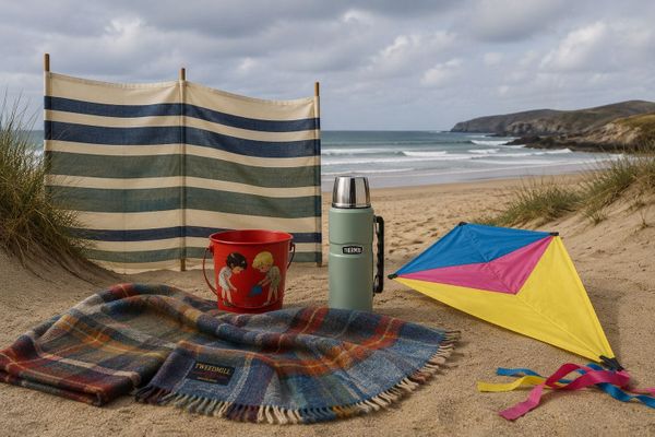 A striped windbreak, a tartan wool picnic rug, a green Thermos flask, a red tin bucket and a colourful kite on a sandy British beach under a grey sky.