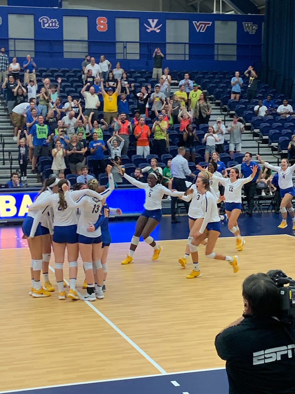 Photo shows an ESPN cameraman capturing the 2019 Pitt Volleyball team and fans celebrating a win over Oregon
