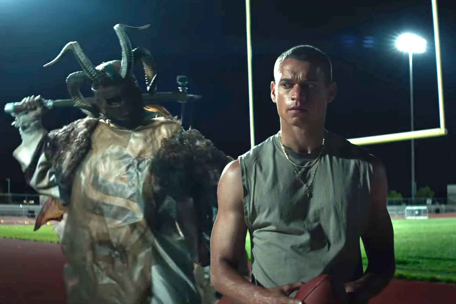 A man holding a football looking over his right shoulder as a larger man looms over him with a hammer.