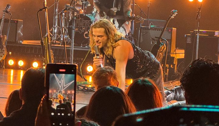 Actor Sam Reid crawling around on the floor of a stage singing at The Vampire Lestat premiere concert. There is a cell phone in the foreground recording.