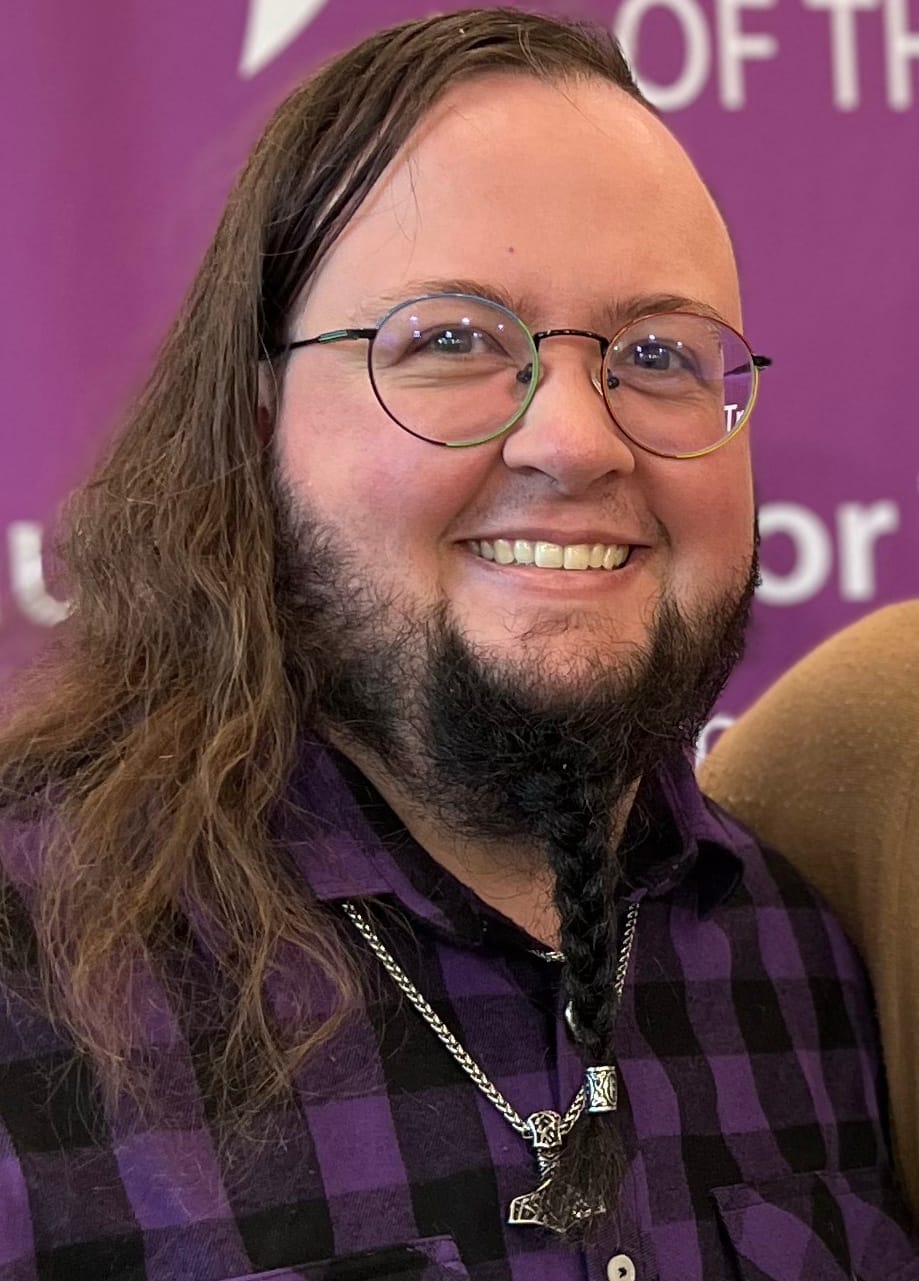 Alyx - a white nonbinary human with shoulder-length brown hair brushed to the side and a dark, braided beard, wearing round glasses and a purple and black plaid shirt - looks directly at the camera