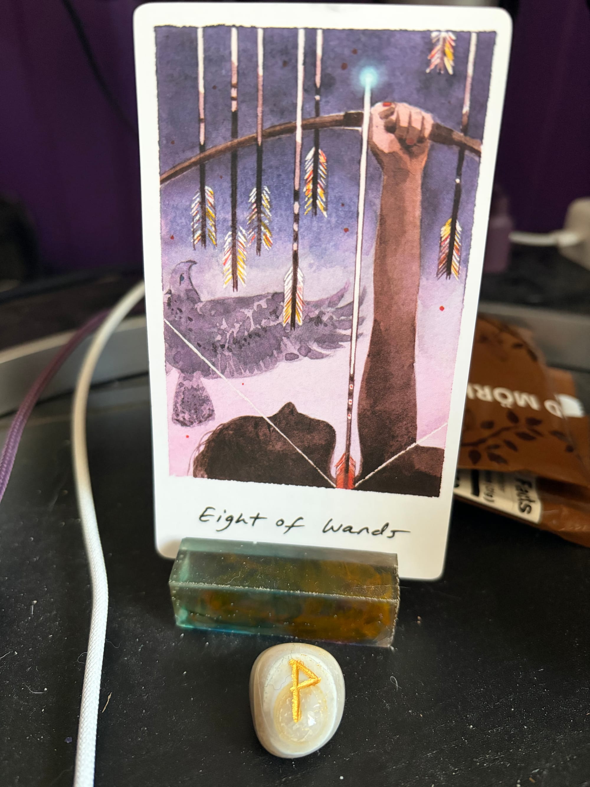 The Eight of Wands from the Yukika Tarot depicts an archer releasing several arrows upward. A bird of prey flies behind the arrows. In front of the card is a small agate with the Wunjo rune carved into it.