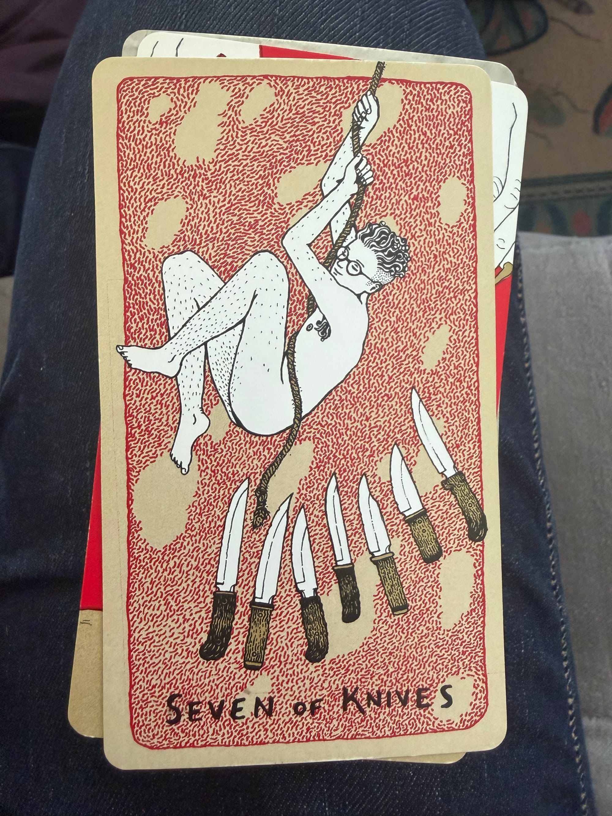 A light-skinned, nude, bespectacled figure with short hair swings on a rope over seven knives with their blades pointed upward