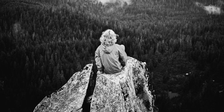 Woman with a raincoat sits with her back to the viewer on a rocky precipice overlooking a forest below with fog through the trees.
