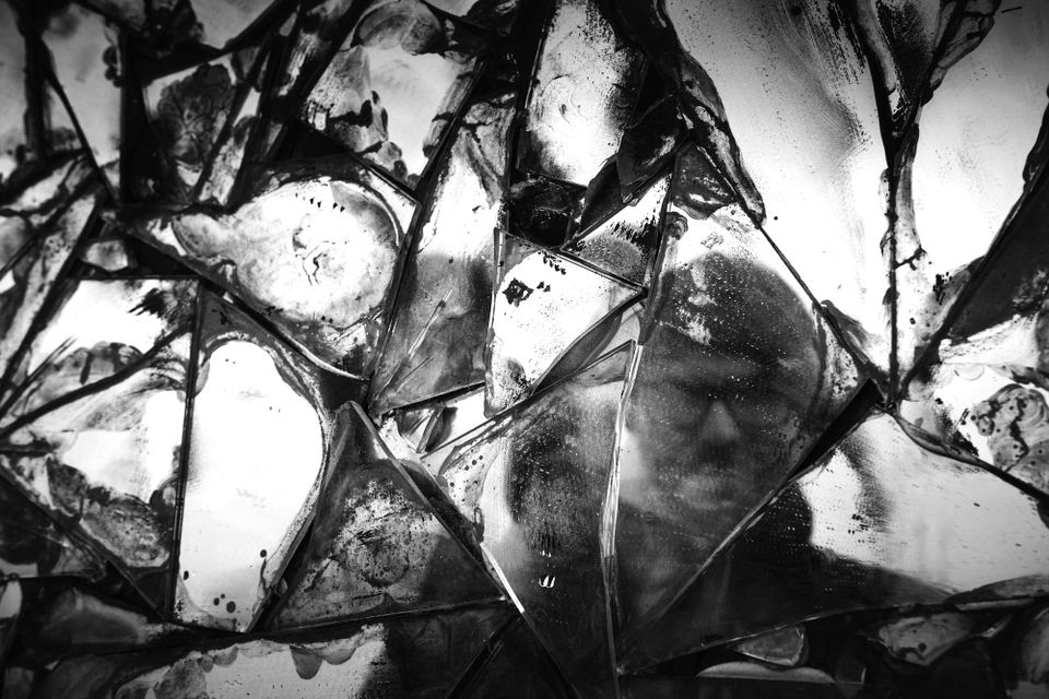 Broken mirrored glass in fragmented chards with a faint reflection of a person with glasses looking through reflection.