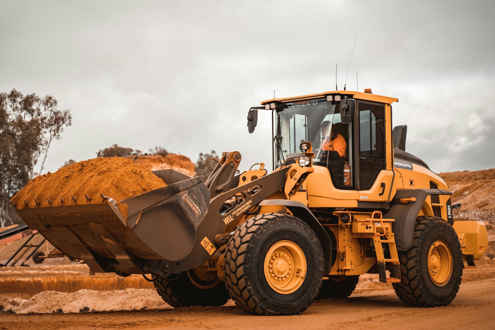 A digger, driven by someone in a high vis, transporting sand on a construction site.