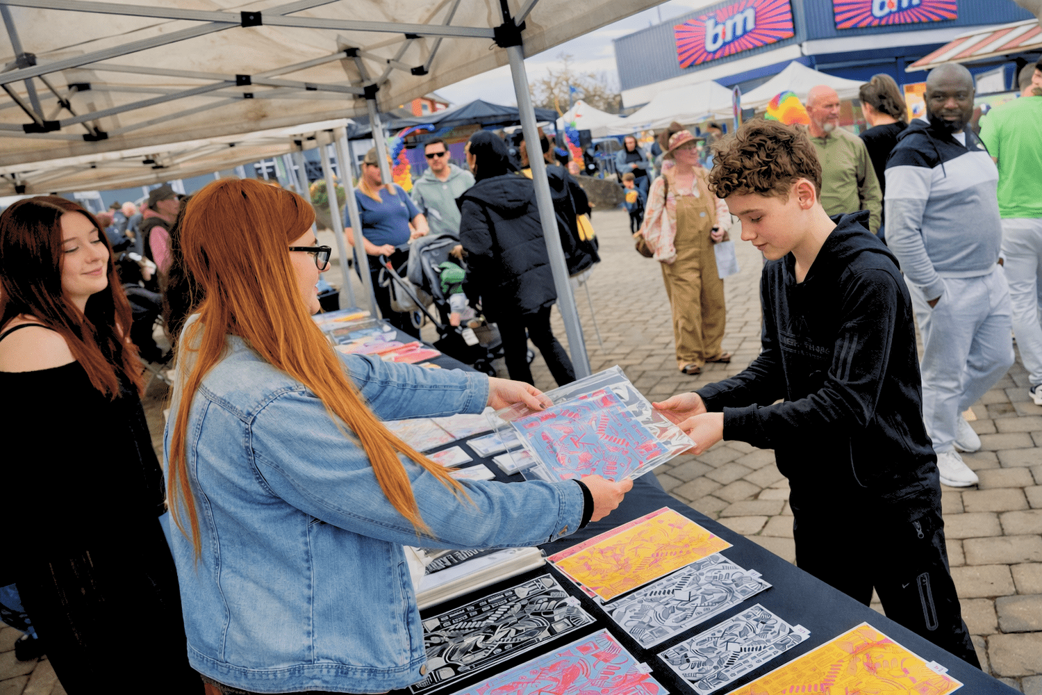 Two teenagers running a market stall, with one handing a piece of art to a customer.