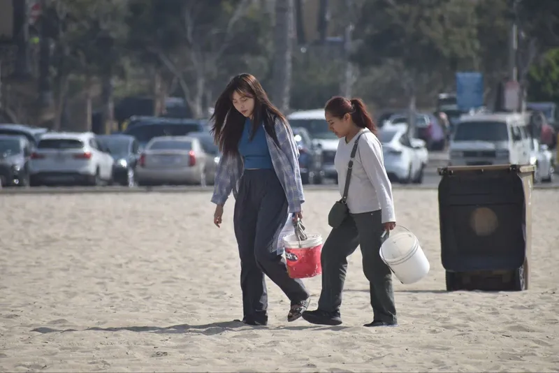Two women walk on a beach with buckets during a beach cleanup day.
