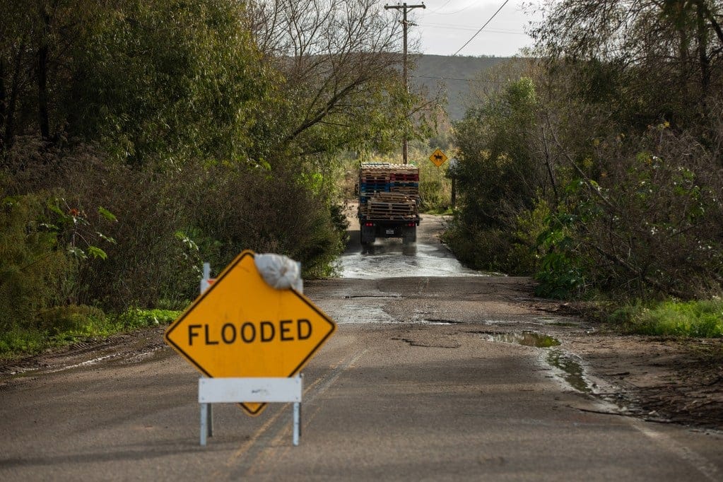 A truck drives by a section of a partially flooded road surrounded by greenery. A yellow caution sign that says "flooded" can be seen in the foreground.