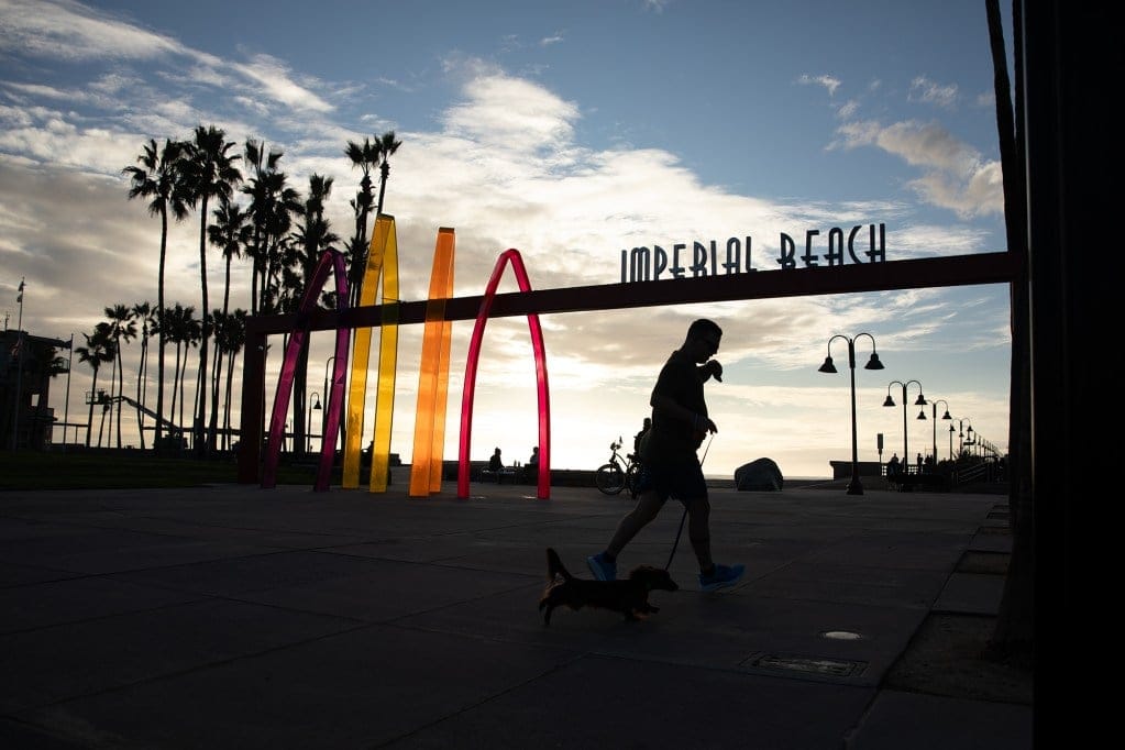 The silhouette of a person walking their dog as they pass by a colorful gateway sign that reads "Imperial Beach" near a beach pier.