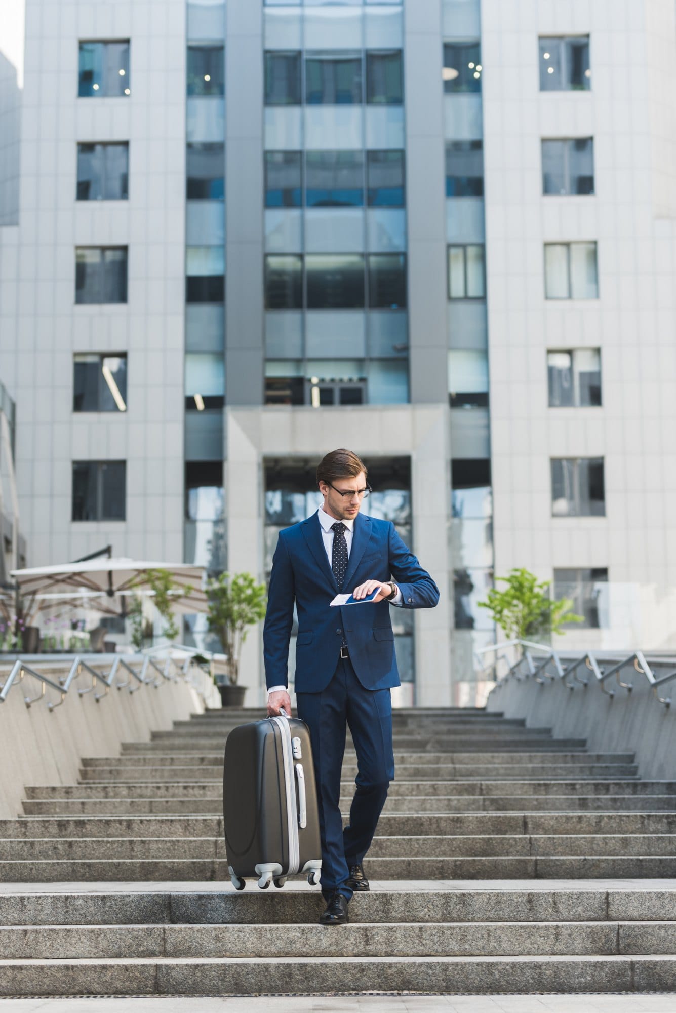 Stylish young businessman with luggage and flight tickets walking by business district and looking