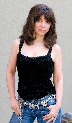 a middle-aged woman with dark hair, wearing blue jeans and a belt of rhinestoned hearts. Her fingers are hooked into her jean pockets and she looks pretty cool