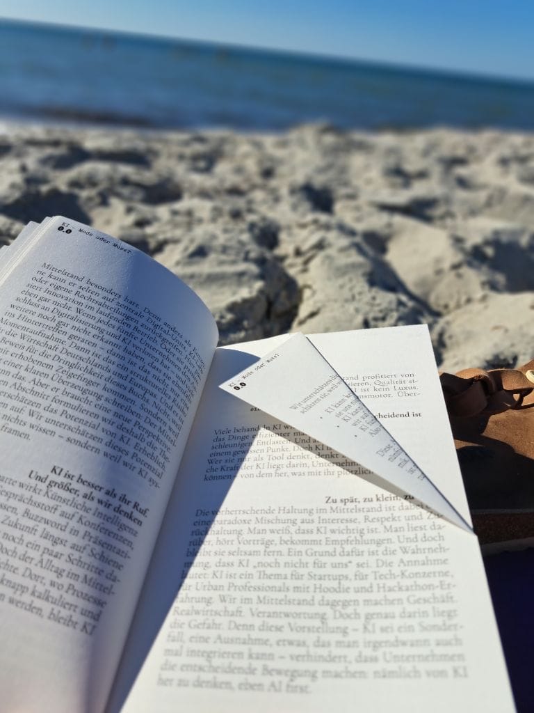 Das Buch "AI first" wird am Strand gelesen. Eine Seite ist als Lesezeichen eingeknickt.