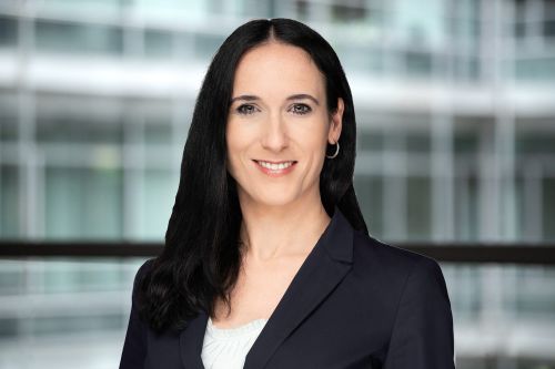 Melanie Kramp-Gerstner im Portrait-Format vor einem verschwommenen Glas-/Stahlhintergrund.