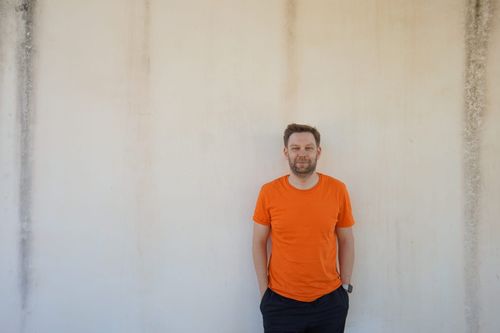 Oliver Voigt steht in einem orangen T-Shirt vor einer Waschbeton-Wand.