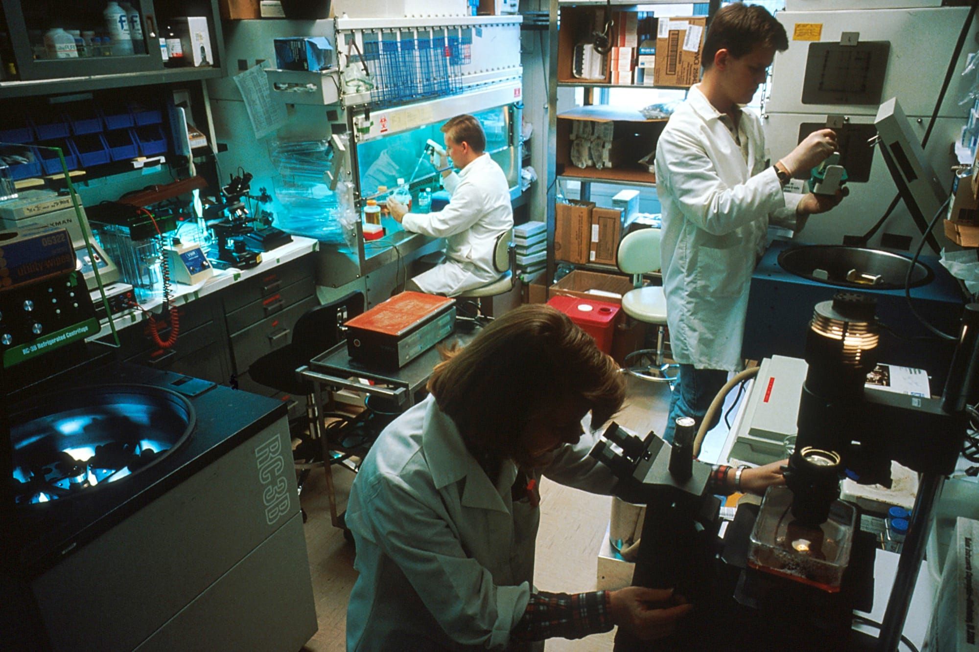 Cercetători în halate albe lucrează la microscoape și hote de laborator