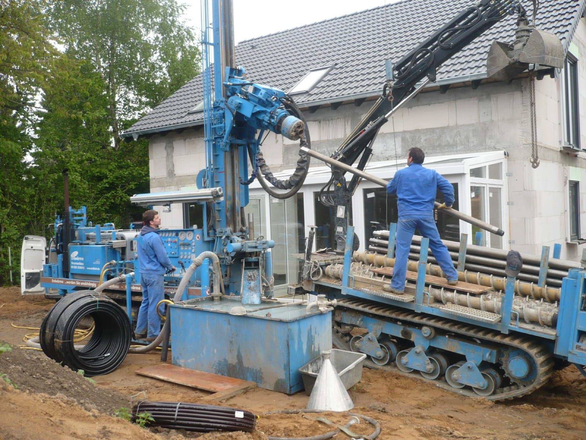 Utilaj de foraj geotermal la o construcție rezidențială, cu doi muncitori și role de țeavă pentru sonde geotermale