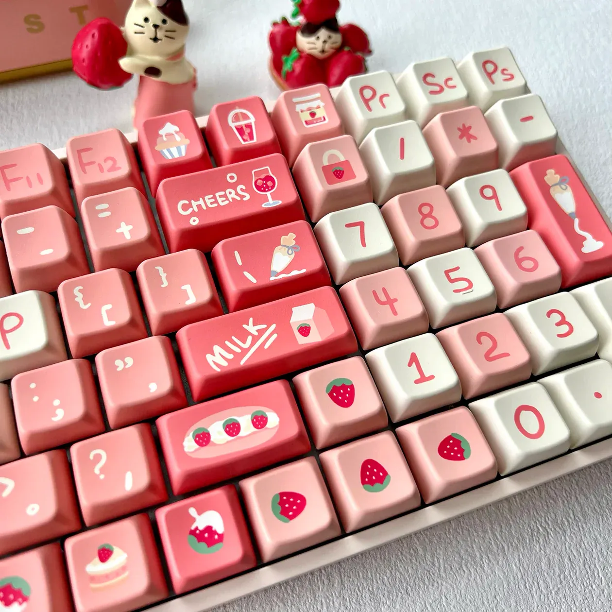A product photo of a strawberry-themed key cap set in pink and white hues with some pink trinkets behind it.