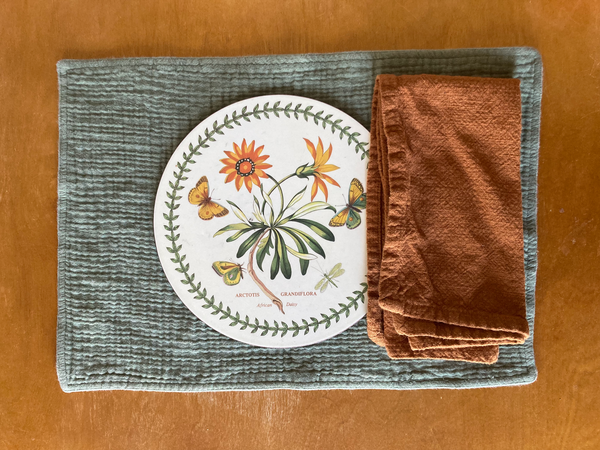 A table setting made of a green placemat, floral charger, and burnt orange cloth napkin sits on top of a wooden tabletop.