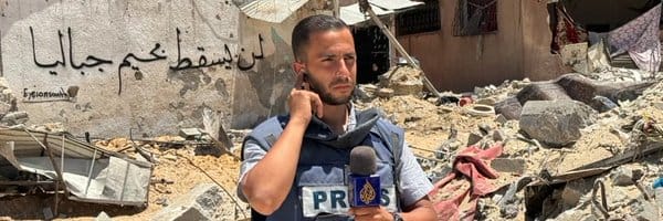 A picture of Anas Al-Sharif in his press vest in front of a bombed site