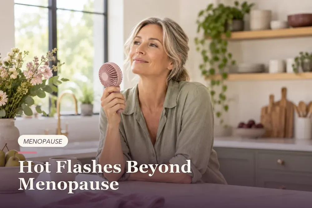 A midlife woman using a handheld fan in a bright kitchen.
