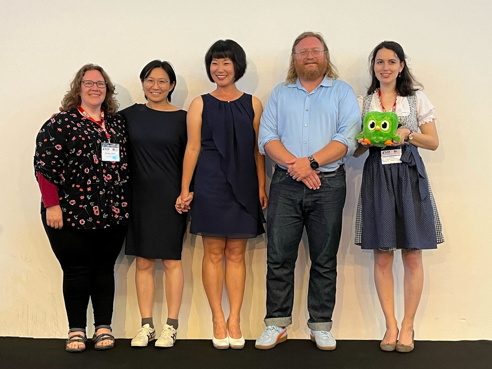 Five people standing and smiling on a stage; the last in line is holding a Duolingo plushie