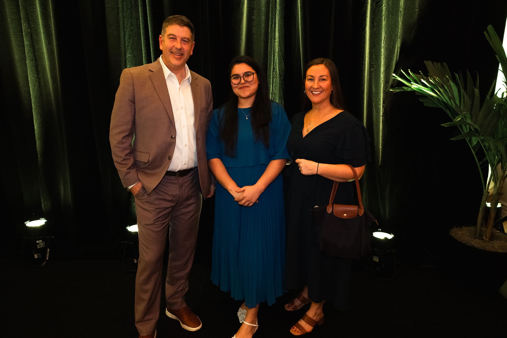 From left, a man and two women stand facing the camera against a green velvet drape. 