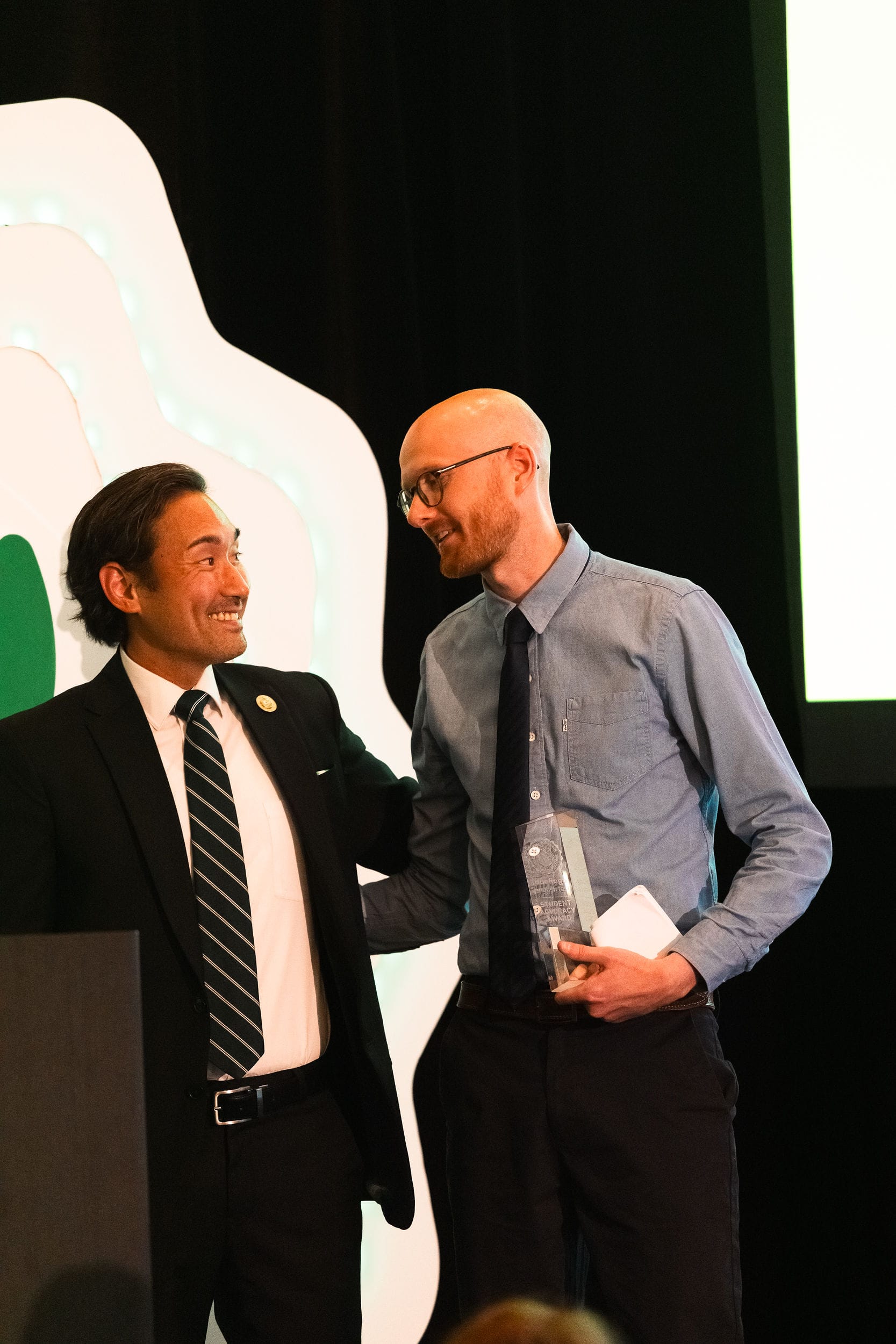 Two man stand together on stage, looking at each other and smiling. The man on the right is holding a We Rise Together award.