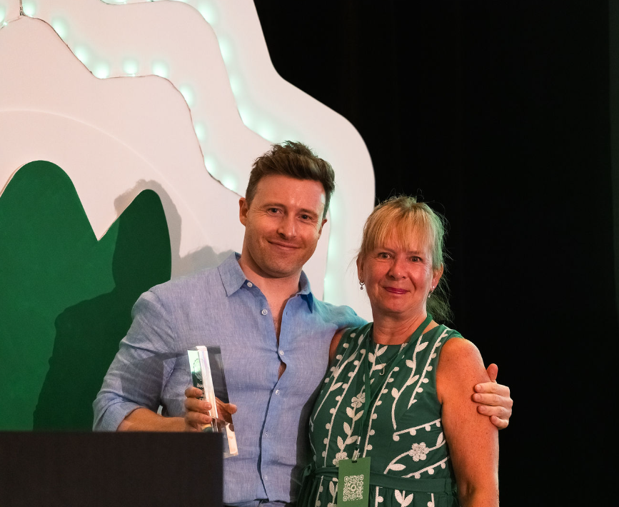 A man and a woman pose on stage, smiling at the crowd. The man is holding an award in his right hand.