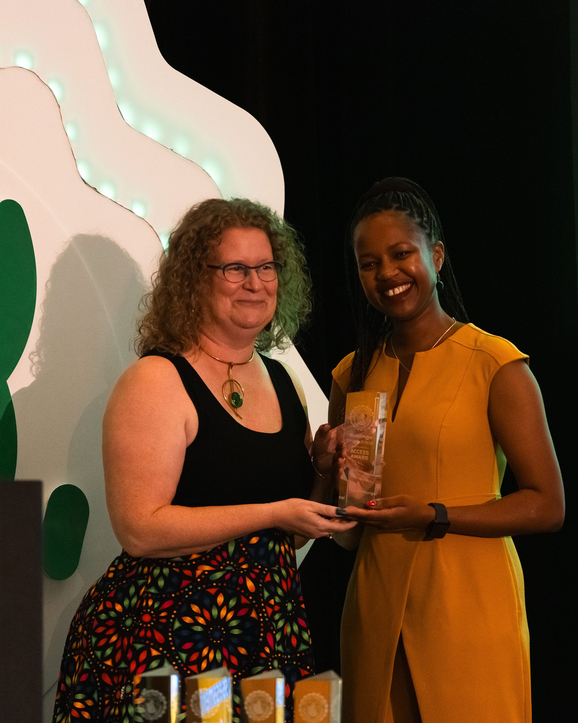 Two women stand together on stage, smiling and facing the crowd. The woman on the left is handing the woman on the right a We Rise Together award