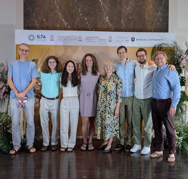 Eight people standing in a line facing the camera. They have their arms around each other and are smiling.