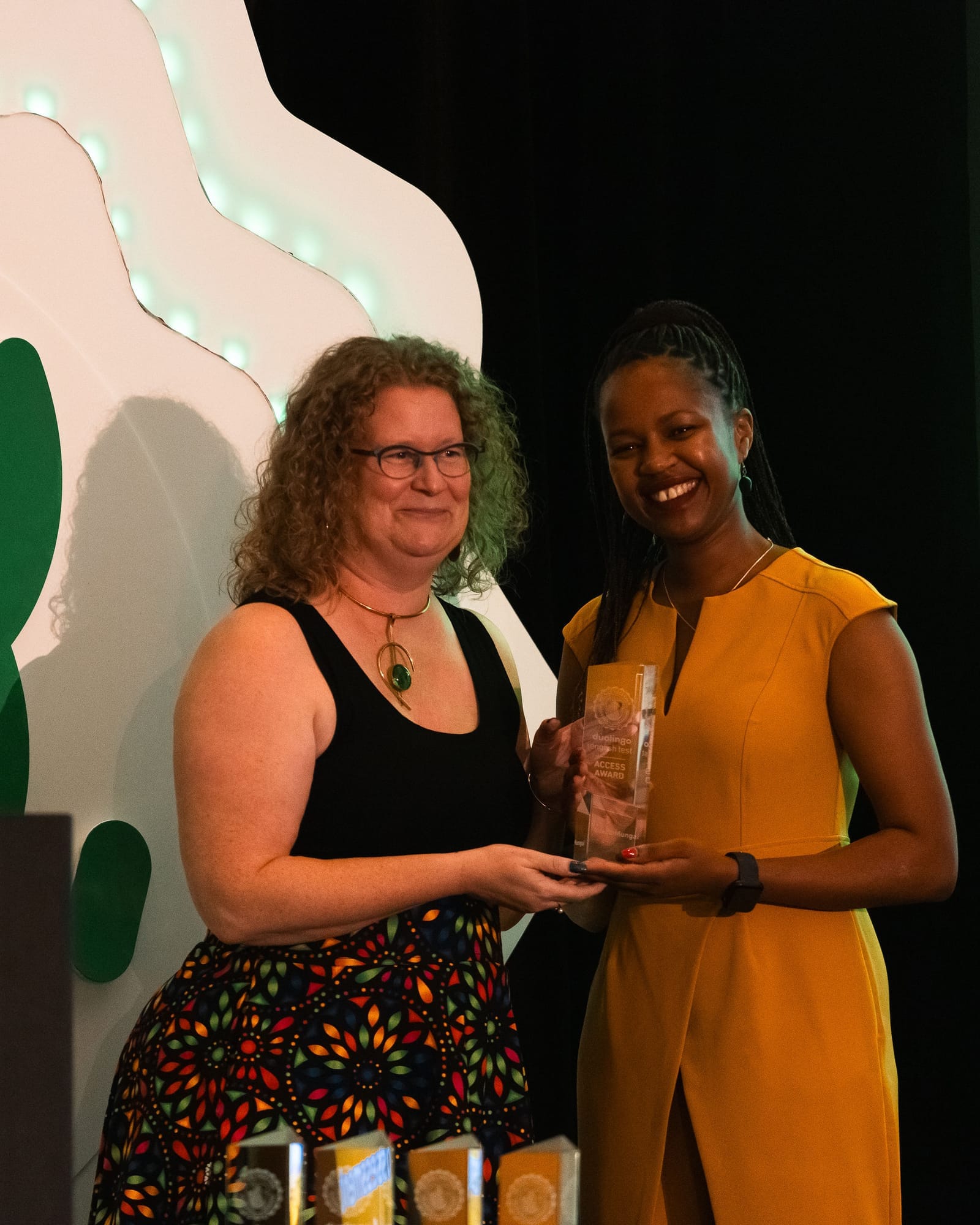 Two women stand together on stage smiling. The woman on the left is handing a clear glass award to the woman on the right.
