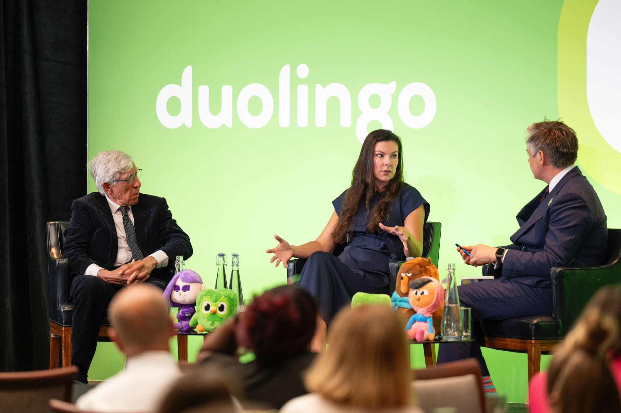 From left, a man, a woman, and a man sit on stage in conversation, in front of a green backdrop that says "duolingo"