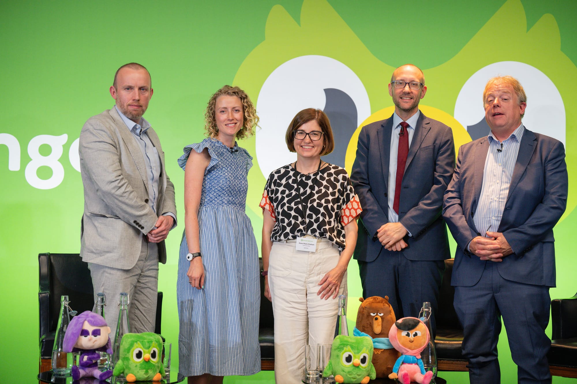 Five people stand on stage in front of a green backdrop with the Duolingo owl. In front of them are plushies of the Duolingo characters.