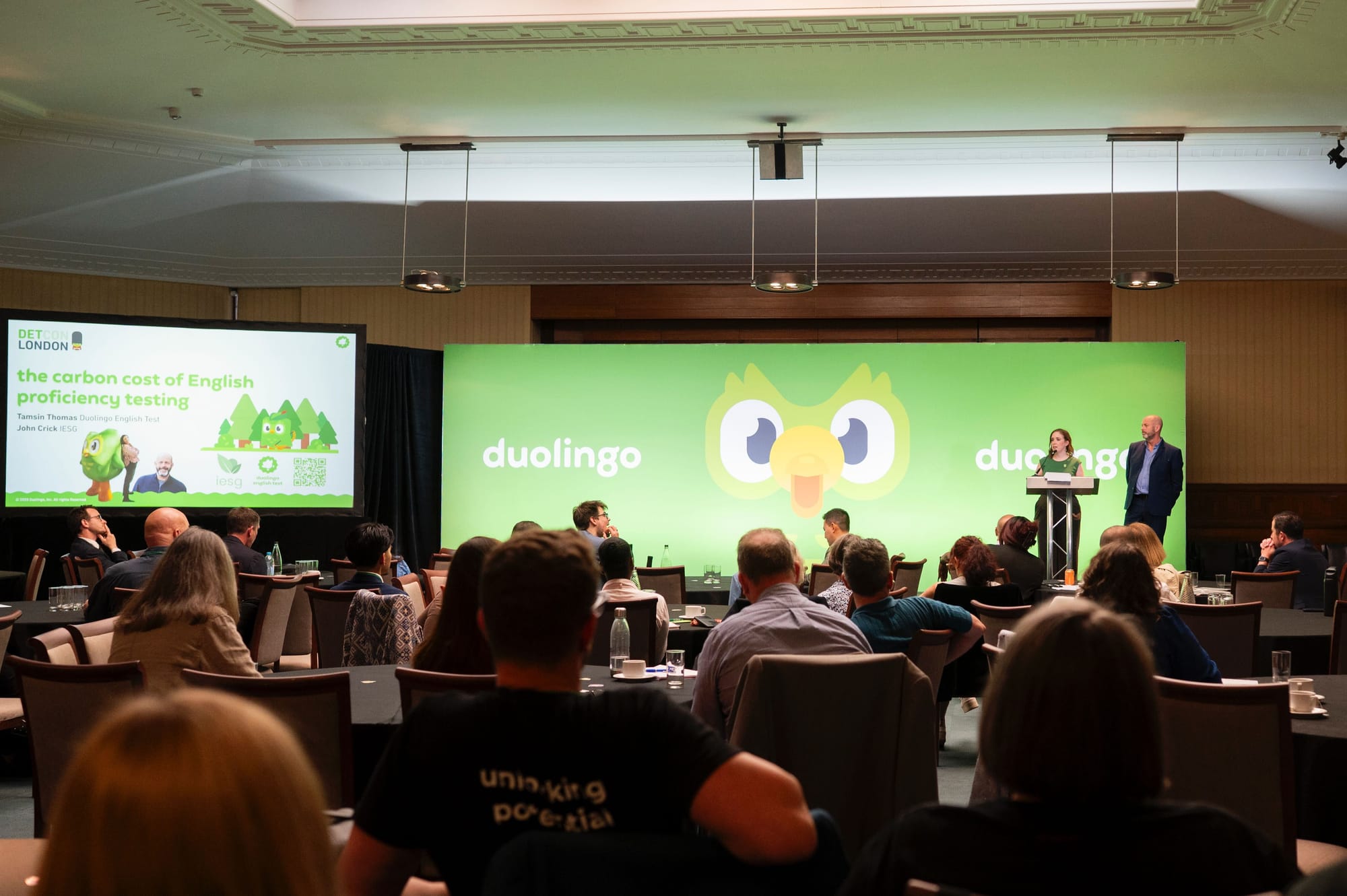 A crowd faces a stage with a green backdrop and the Duolingo owl. Two people on stage are presenting.