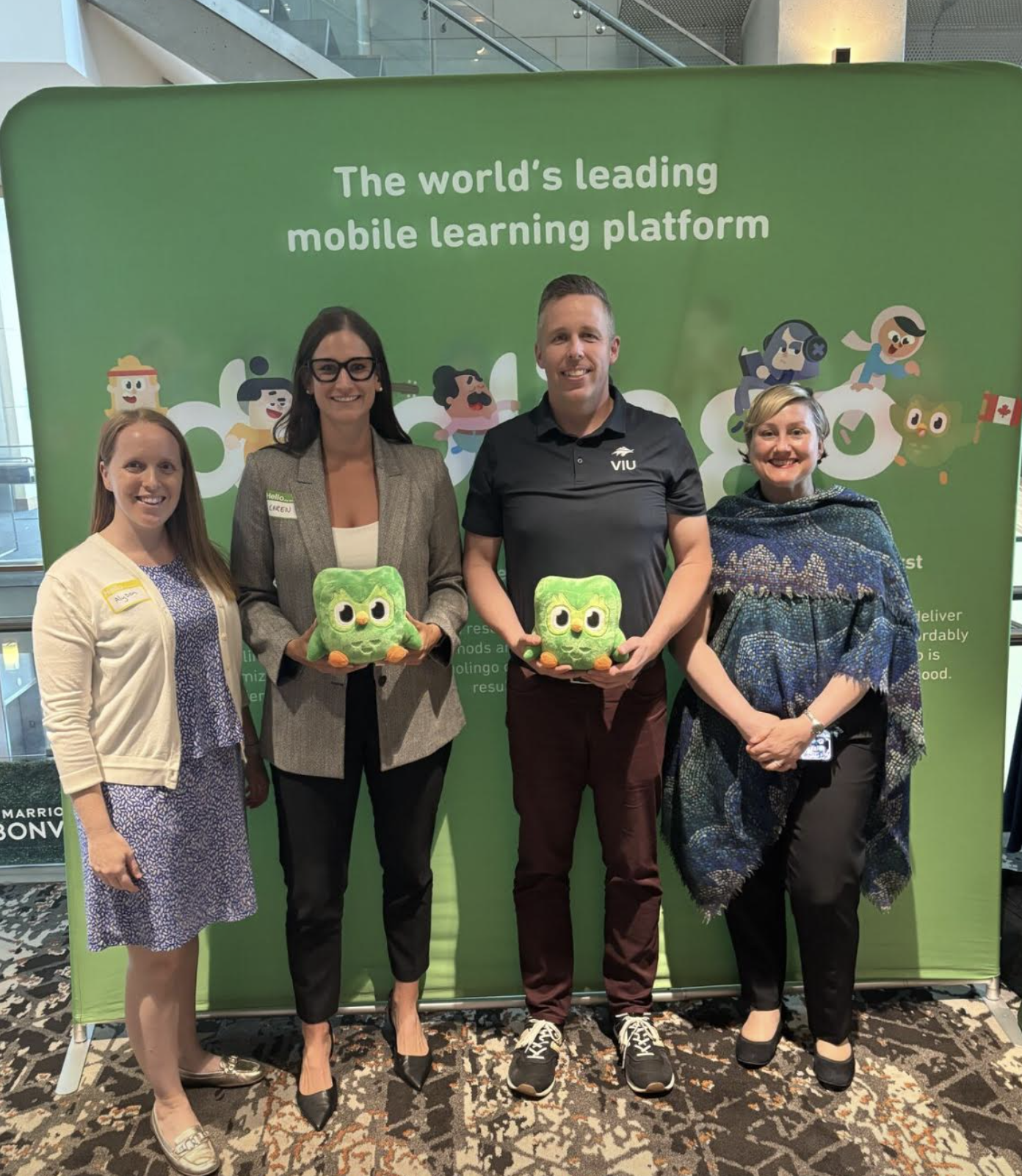 Four people stand in a line against a green duolingo backdrop, smiling. The two people in the middle are holding plushies of Duo, the Duolingo Owl. 