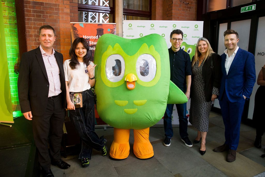 a man in a suit, a woman in a white t-shirt, a green owl mascot, a man in a black shirt, a woman in a dotted ress, and a man in a suit face the camera