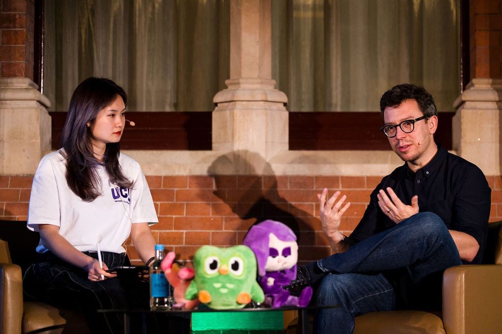 On the left, a woman in a white shirt sits in a chair, on the right, a man in a black shirt sits in a chair, speaking to the crowd.