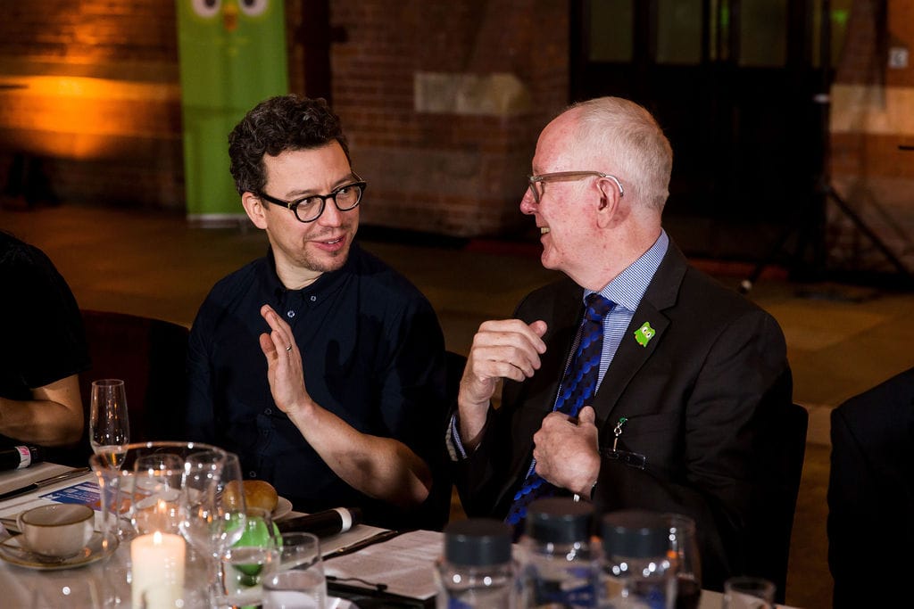Two men sit at a banquet table. They are turned towards each other and are smiling, in mid conversation.